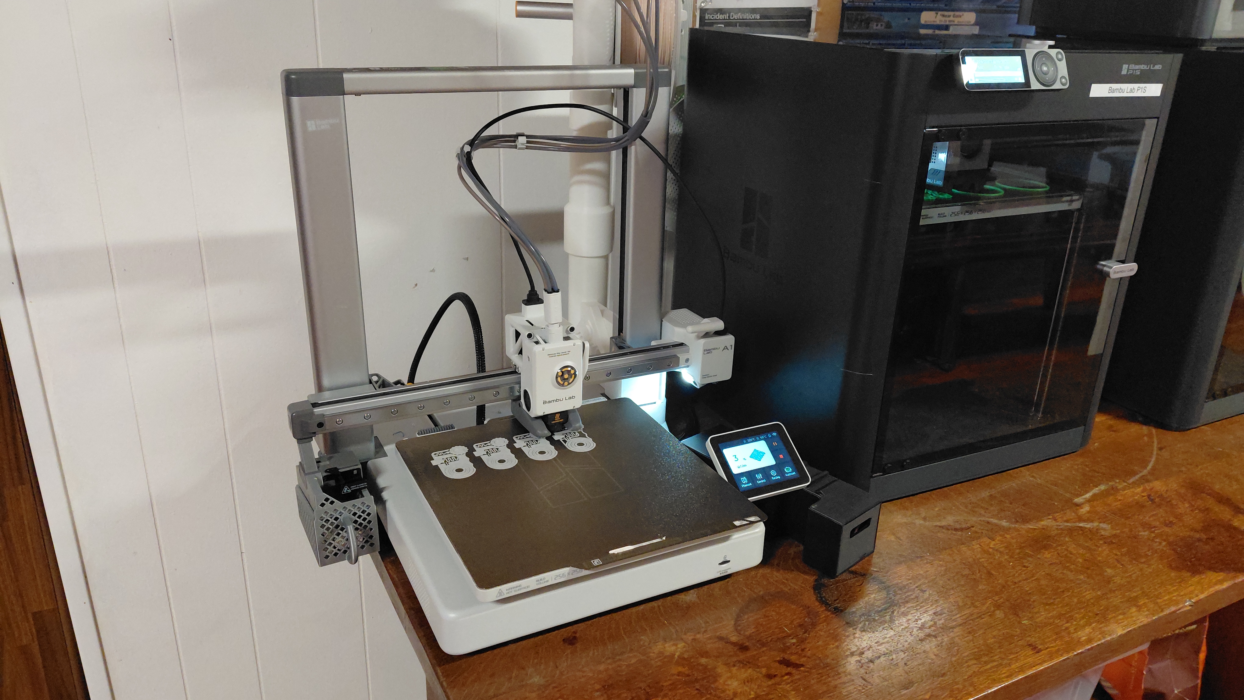 Two Bambu Lab 3D printers on a wooden bench, one printing small parts