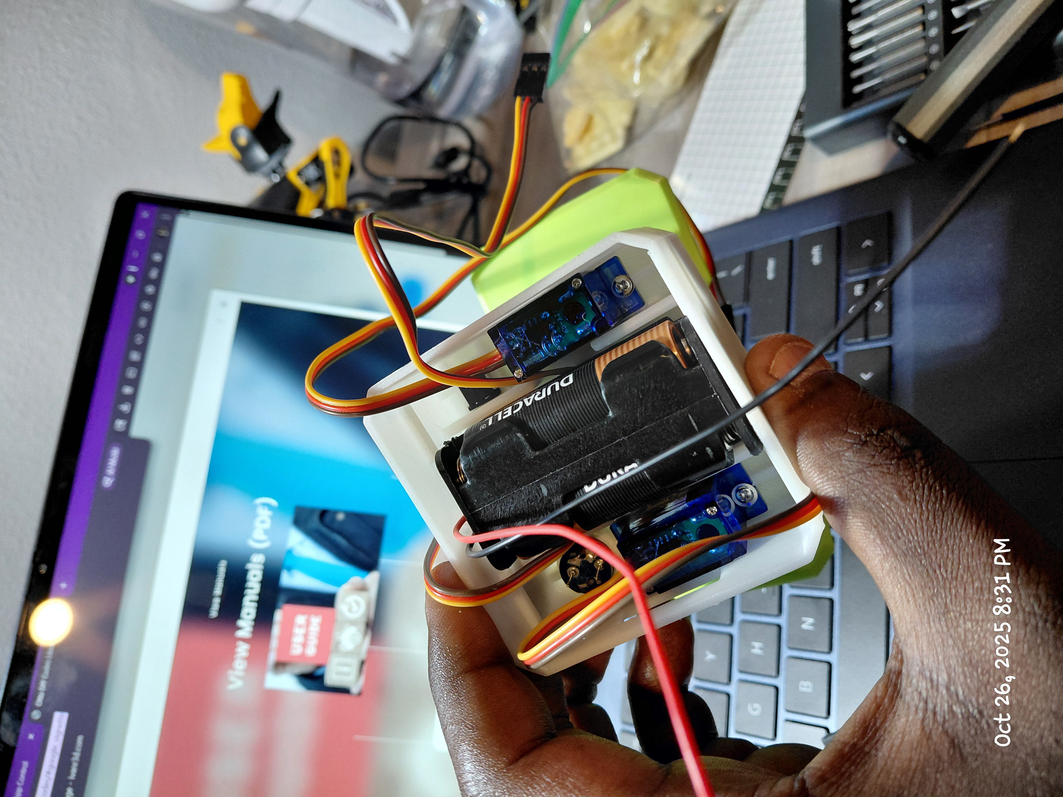 Hand holding a small 3D-printed robot chassis with servos and battery holder near a laptop